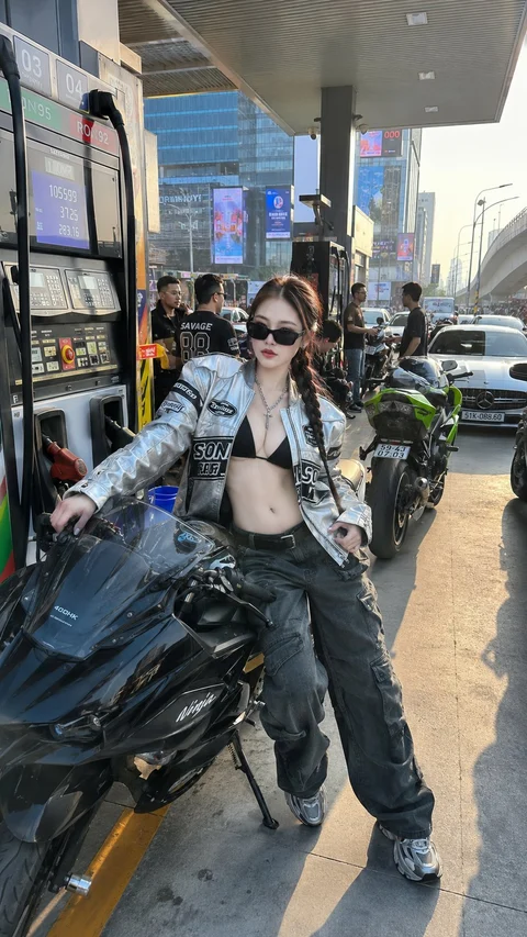 A woman in sunglasses, a patterned jacket and dark trousers poses beside a motorcycle at a roadside stop.